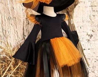 May include: A black and orange Halloween costume for children. The costume includes a black dress with bell sleeves, an orange tulle skirt, and a matching witch hat with an orange bow. The outfit is displayed on a mannequin.
