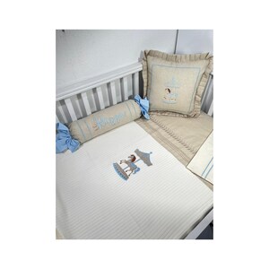 May include: A baby crib set featuring a white mattress with a carousel horse embroidery. Includes a bolster pillow with the name "Razgoyon" embroidered in blue, a square pillow, and a beige blanket. The set has blue accents and is designed for a nursery.