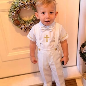 May include: A white short-sleeved shirt and trousers set with suspenders and a bow tie. The shirt features a gold embroidered cross on the chest, the name Adam on the right sleeve, and the date 06.22.25 on the left sleeve. White sneakers complete the outfit.