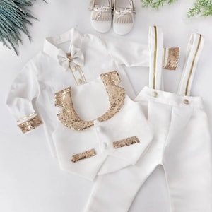 May include: A white formal outfit for a baby, including a long-sleeved shirt with a bow tie, a vest with gold sequins, pants with suspenders, and matching shoes. The outfit has gold accents.