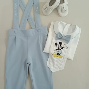 May include: A light blue baby outfit with suspenders, a white shirt with a bow tie and a Mickey Mouse design, and white shoes with tassels. The suspenders have the name "Kuzey" and the number "1" embroidered on them.