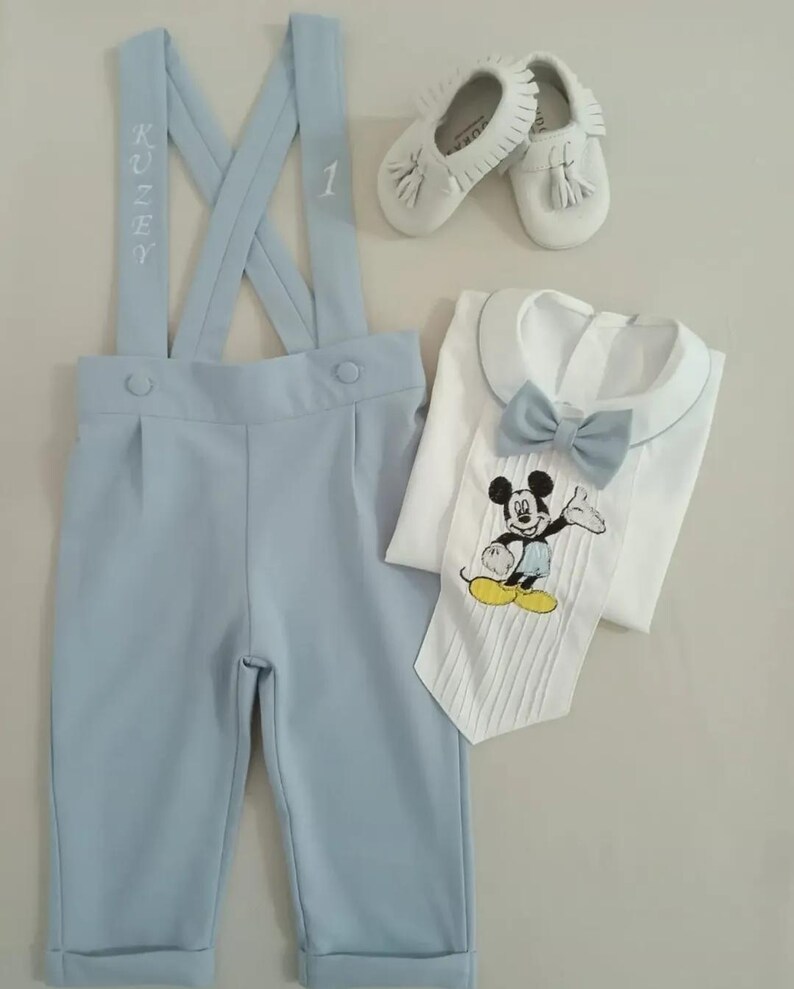 May include: A light blue baby outfit with suspenders, a white shirt with a bow tie and a Mickey Mouse design, and white shoes with tassels. The suspenders have the name "Kuzey" and the number "1" embroidered on them.