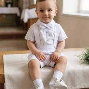Puede incluir: Un conjunto blanco de camisa de manga corta con pajarita, tirantes y pantalón corto. La camisa presenta una cruz bordada en plata en el pecho y el nombre Marion con la fecha 20.06.2024 en las mangas. Calcetines y zapatos blancos completan el conjunto.