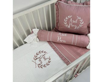 Può includere: Un lettino bianco con un set di biancheria da letto rosa e bianco. La biancheria da letto ha un motivo a righe bianche e rosa e presenta un cuscino rosa con il nome "Asel Pera" ricamato su di esso. Il lettino ha anche un cuscino rosa e bianco con un design a corona e il nome "Asel Pera" ricamato su di esso.