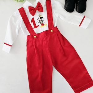 May include: A white button-down shirt with red trim and a red bow tie. The shirt features a Mickey Mouse character wearing a crown and the number "1" embroidered on the front. The shirt is paired with red suspenders and red pants.
