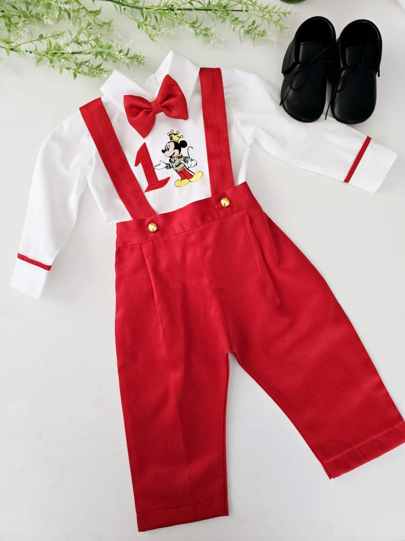 May include: A white button-down shirt with red trim and a red bow tie. The shirt features a Mickey Mouse character wearing a crown and the number "1" embroidered on the front. The shirt is paired with red suspenders and red pants.