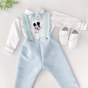 May include: A baby boy's outfit with a white shirt, blue pants, and a bow tie. The shirt has a Mickey Mouse embroidery and the pants have suspenders. The outfit is personalized with the name "Franklin".