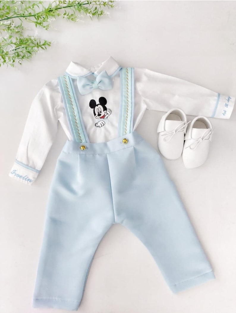 May include: A baby boy's outfit with a white shirt, blue pants, and a bow tie. The shirt has a Mickey Mouse embroidery and the pants have suspenders. The outfit is personalized with the name "Franklin".