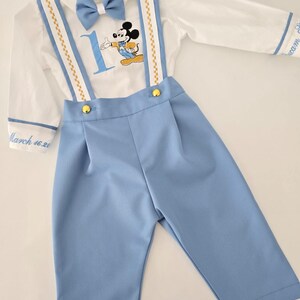 May include: A white long-sleeve shirt with a blue bow tie, gold suspenders, and a Mickey Mouse graphic. The shirt has the number "1" and the text "March 16, 20" embroidered on it. The shirt is paired with blue pants.