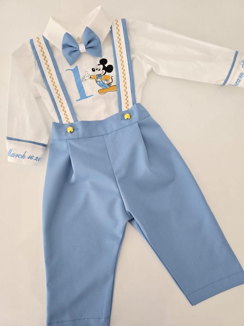 May include: A white long-sleeve shirt with a blue bow tie, gold suspenders, and a Mickey Mouse graphic. The shirt has the number "1" and the text "March 16, 20" embroidered on it. The shirt is paired with blue pants.