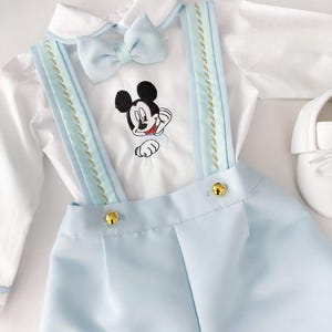 May include: A baby boy's outfit with a white shirt, blue overalls, and a bow tie. The overalls have gold buttons and a Mickey Mouse design embroidered on the shirt.