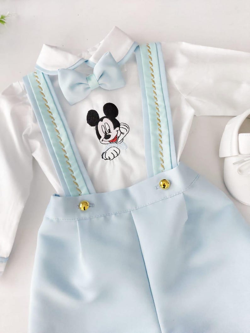May include: A baby boy's outfit with a white shirt, blue overalls, and a bow tie. The overalls have gold buttons and a Mickey Mouse design embroidered on the shirt.