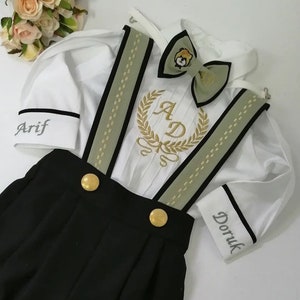 Pode incluir: Uma camisa branca de botões com acabamento preto e uma gravata borboleta verde. A camisa tem as letras "AD" bordadas na frente com uma coroa dourada. A camisa é combinada com calças pretas com botões dourados e suspensórios verdes. A camisa tem os nomes "Arif" e "Doruk" bordados nas mangas.