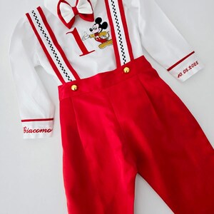 May include: A white button-down shirt with red trim and a Mickey Mouse graphic. The shirt has a bow tie and is worn with red overalls with black and white checkered straps. The overalls have gold buttons and the text "Giacomo" and "40.05.2025" is embroidered on the garment.