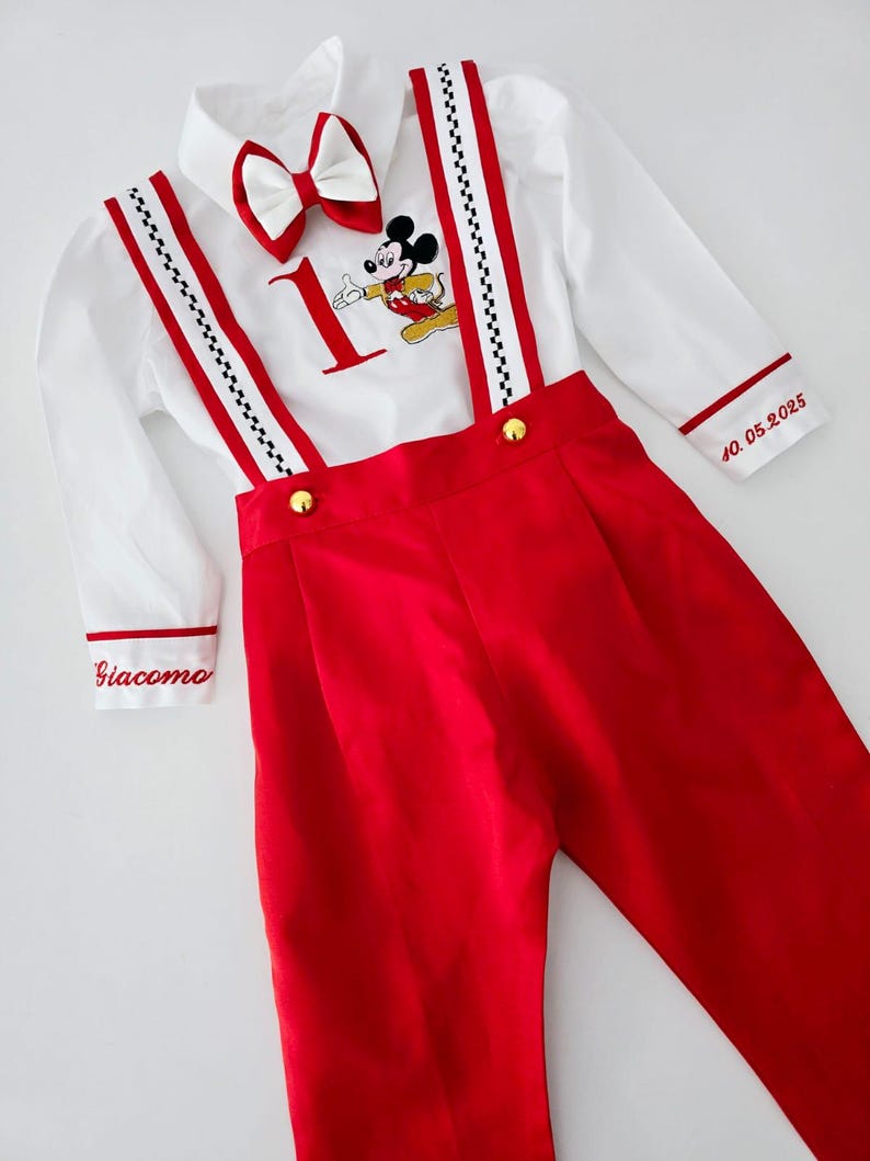 May include: A white button-down shirt with red trim and a Mickey Mouse graphic. The shirt has a bow tie and is worn with red overalls with black and white checkered straps. The overalls have gold buttons and the text "Giacomo" and "40.05.2025" is embroidered on the garment.