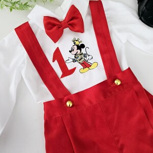 May include: A white button-down shirt with a red bow tie and red suspenders. The shirt has an embroidered image of Mickey Mouse wearing a crown and holding a number one. The suspenders have gold buttons.