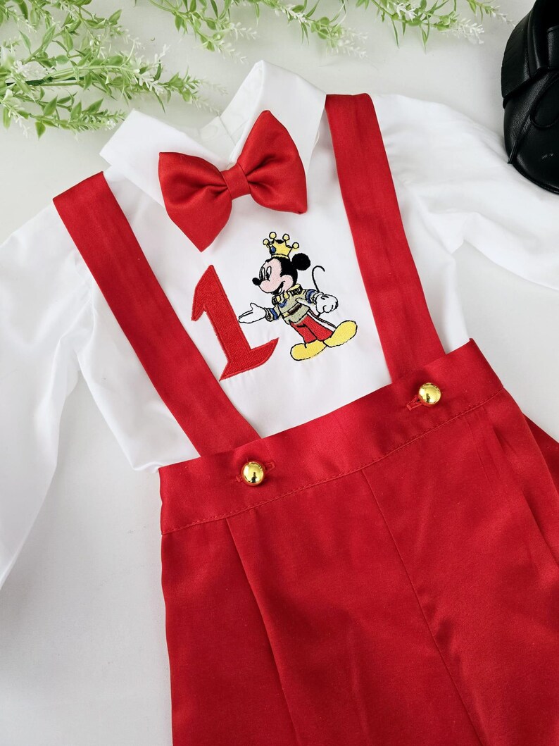 May include: A white button-down shirt with a red bow tie and red suspenders. The shirt has an embroidered image of Mickey Mouse wearing a crown and holding a number one. The suspenders have gold buttons.