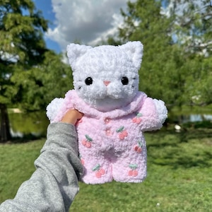 K&ouml;nnte beinhalten: Wei&szlig;es geh&auml;keltes Katzen-Pl&uuml;schtier, das einen rosa Schlafanzug mit Kirschmuster tr&auml;gt.