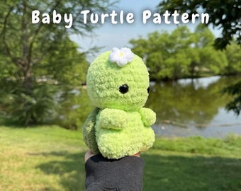 May include: A handmade, light green crochet baby turtle with a white flower on its head. The turtle has black eyes and is held up against a background of green trees and a lake. The text "Baby Turtle Pattern" is displayed above the turtle.