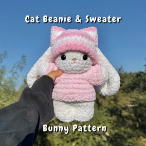 Op de afbeelding: Een handgemaakte gehaakte konijn met een roze en wit gestreepte kattenmuts en een roze trui. Het konijn heeft lange witte oren en zwarte ogen. De tekst "Cat Beanie & Sweater" en "Bunny Pattern" zijn zichtbaar.