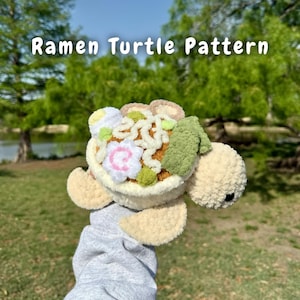 Puede incluir: Un juguete de peluche de tortuga de crochet con un tazón de ramen en la espalda. El tazón de ramen está lleno de fideos, algas marinas y una torta de pescado rosa. La tortuga es beige y el tazón de ramen es marrón.