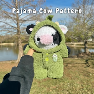 Puede incluir: Un juguete de peluche de vaca con un pijama verde con capucha. La vaca tiene manchas blancas y negras y una nariz rosa. El pijama tiene un botón verde y una bebida verde con una pajita en la parte delantera. El texto "Pajama Cow Pattern" está en la parte superior de la imagen.