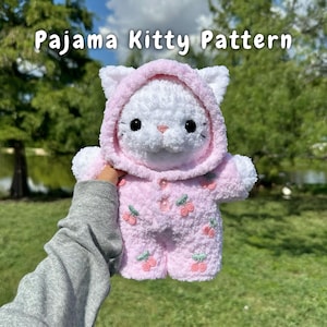 Könnte beinhalten: Ein weißes gehäkeltes Kätzchen trägt einen rosa Kapuzenpyjama mit einem Kirschmuster. Der Pyjama hat einen Knopfverschluss und ist mit grünen Blättern und rosa Kirschen bestickt.
