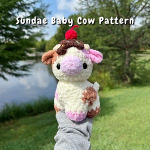 Puede incluir: Un peluche de vaca color crema con manchas marrones y rosas, un hocico rosa y ojos negros. La vaca está coronada con un helado de chocolate, chispas y una cereza roja. El texto "Sundae Baby Cow Pattern" está encima.