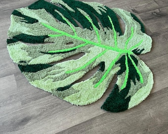 May include: A green and white monstera leaf shaped rug. The rug is made of a soft, plush material and has a realistic leaf design.