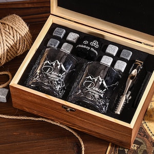 Könnte beinhalten: Geöffnete Holzgeschenkbox mit zwei gravierten Whiskygläsern, Whisky-Steinen und einer Metall-Eiszange. Die Gläser sind mit einem Bergmotiv und dem Namen "Daniel" graviert.