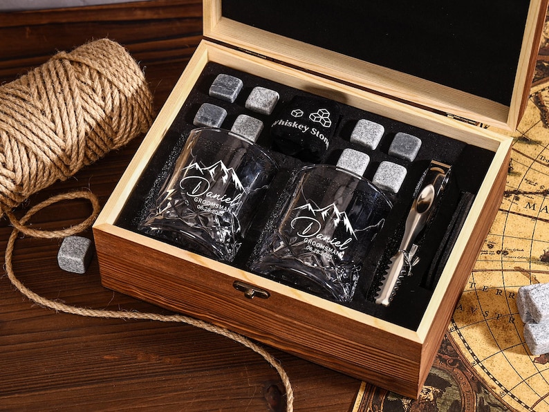 May include: A wooden gift box with two engraved whiskey glasses, whiskey stones, and a metal ice tong. The glasses are etched with a mountain design and the name "Daniel". The box is open, revealing the contents.