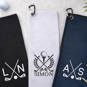 Könnte beinhalten: Drei Golfhandtücher in Schwarz, Weiß und Marineblau, jedes mit einem schwarzen Karabinerhaken. Die Handtücher zeigen Golfmotive, darunter gekreuzte Golfschläger, Golfbälle und personalisierte Initialen oder Namen. Golfzubehör.