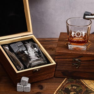 May include: A personalized whiskey glass with the name "SIMON" and a monogram, resting on a wooden box. The box contains whiskey stones, a glass, and a metal ice tong. The glass is filled with amber liquid. The set is presented on a wooden surface.