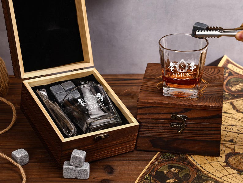 May include: A personalized whiskey glass with the name "SIMON" and a monogram, resting on a wooden box. The box contains whiskey stones, a glass, and a metal ice tong. The glass is filled with amber liquid. The set is presented on a wooden surface.