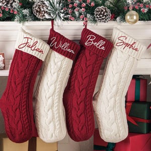 May include: Four cable-knit Christmas stockings in red and cream, personalized with names: Julian, William, Bella, and Sophia. The stockings are hanging on a white mantelpiece, decorated with festive greenery and ornaments.