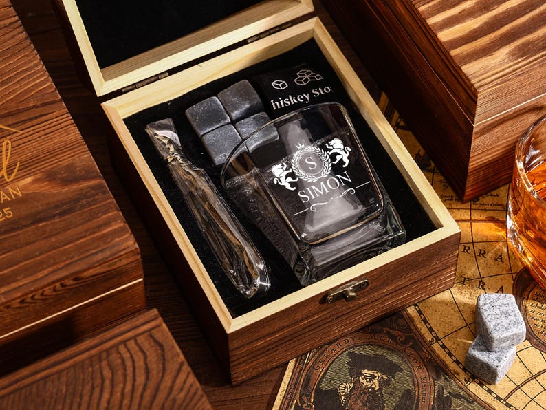 May include: A wooden gift box set containing a rocks glass with "SIMON" printed on it, whiskey stones, and a stainless steel ice wand. The box is open, revealing the contents on a black velvet lining.