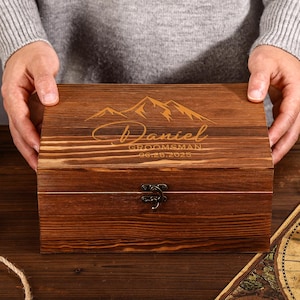 May include: A wooden box with a dark brown finish, featuring engraved mountain imagery and the name "Daniel" with the text "Groomsman 06.28.2025". The box has a metal clasp and is held by a person.