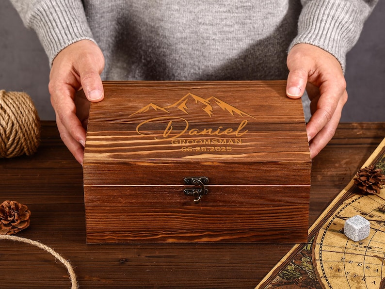 May include: A wooden box with a dark brown finish, featuring engraved mountain imagery and the name "Daniel" with the text "Groomsman 06.28.2025". The box has a metal clasp and is held by a person.