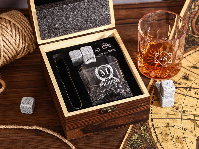 May include: A wooden gift box set with a whiskey glass, ice cubes, and tongs. The glass is engraved with a monogram and the name "MICHAEL". A second glass with initials "JS" is also visible. The set includes whiskey stones.