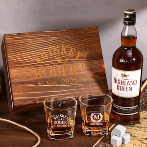May include: A whiskey gift set featuring a wooden box with "Whiskey Robert Est. 2025" engraved on it, two shot glasses, a bottle of Highland Queen Blended Scotch Whisky, and whiskey stones. The set is arranged on a wooden surface.