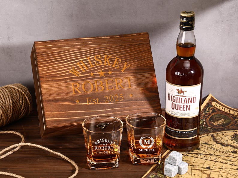 May include: A whiskey gift set featuring a wooden box with "Whiskey Robert Est. 2025" engraved on it, two shot glasses, a bottle of Highland Queen Blended Scotch Whisky, and whiskey stones. The set is arranged on a wooden surface.