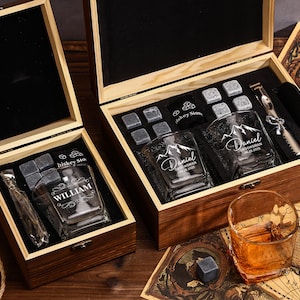 May include: Two wooden gift boxes containing whiskey glasses, ice stones, and a metal tool. One box has a glass engraved with "William". The other box has two glasses engraved with "Daniel". The glasses are clear, and the boxes are brown.