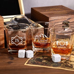 May include: Three personalized whiskey glasses with ice cubes, filled with amber liquid, sit on a wooden surface. Each glass features a different design and name. A wooden box is in the background.