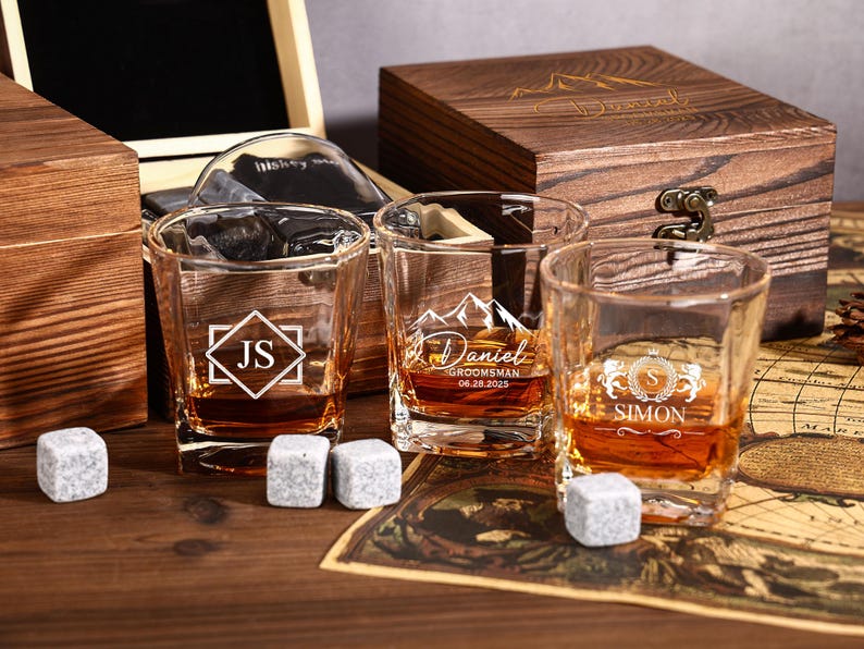 May include: Three personalized whiskey glasses with ice cubes, filled with amber liquid, sit on a wooden surface. Each glass features a different design and name. A wooden box is in the background.