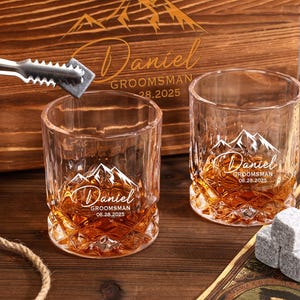 May include: Two crystal whiskey glasses with a mountain design and the name "Daniel" engraved. The glasses are filled with amber liquid and sit on a wooden surface. A wooden box with matching engraving is in the background. Ice cubes and tongs are also present.