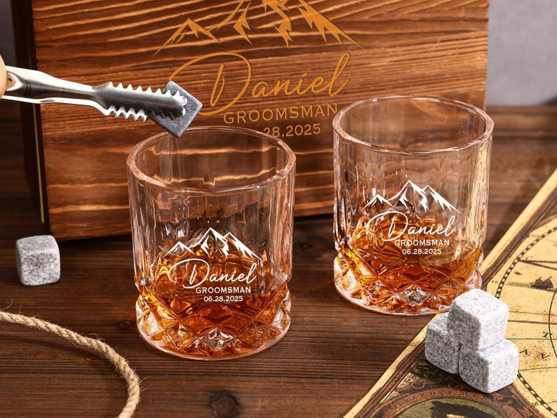 May include: Two crystal whiskey glasses with a mountain design and the name "Daniel" engraved. The glasses are filled with amber liquid and sit on a wooden surface. A wooden box with matching engraving is in the background. Ice cubes and tongs are also present.