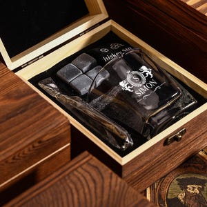 May include: A wooden gift box with a glass tumbler, whiskey stones, and a glass straw. The tumbler is engraved with the name "SIMON" and a crest. The box is made of dark wood.