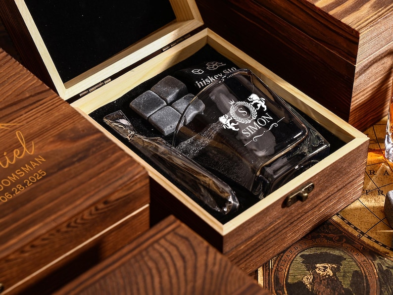 May include: A wooden gift box with a glass tumbler, whiskey stones, and a glass straw. The tumbler is engraved with the name "SIMON" and a crest. The box is made of dark wood.