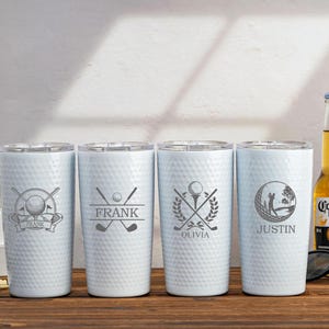 Könnte beinhalten: Vier weiße Golf-Tumbler mit personalisierten Namen: Frank, Olivia, Justin. Jeder Tumbler zeigt ein anderes Golf-Design. Die Tumbler stehen auf einer Holzoberfläche.