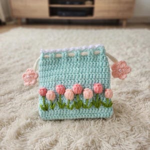 Può includere: Un astuccio all'uncinetto azzurro, decorato con una fila di tulipani rosa e steli verdi. Il bordo superiore presenta un bordo bianco e crema, con accenti floreali rosa sui cordini. L'astuccio è posto su uno sfondo morbido e bianco.